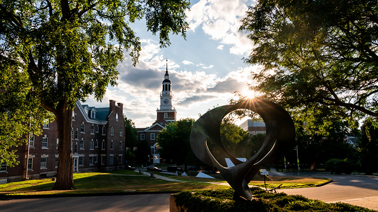 Photo of Baker Tower through trees with a circular sculpture in the foreground and sunbeams hitting the sculpture through the trees