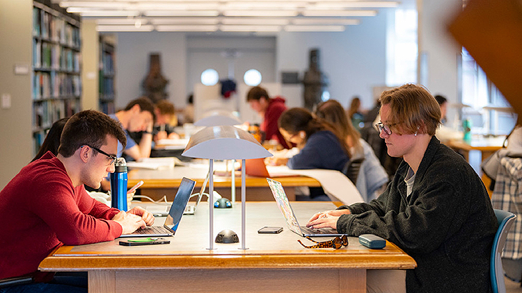 Many students sit at tables in the library studying or on their computers