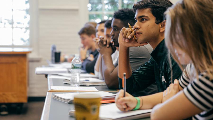 A line of students sit in class taking notes and listening to the professor