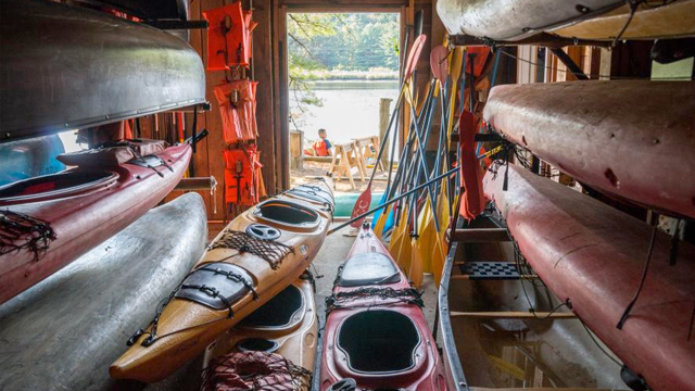 Kayaks and canoes in the foreground inside the boathouse as students gather by the Connecticut River