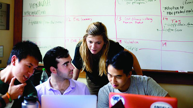 A woman standing behind three men, looking at a computer screen and engaging in conversation. 