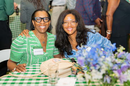 Two guests seated at a table posing and smiling. The table has a floral arrangement and a green and white tablecloth. 