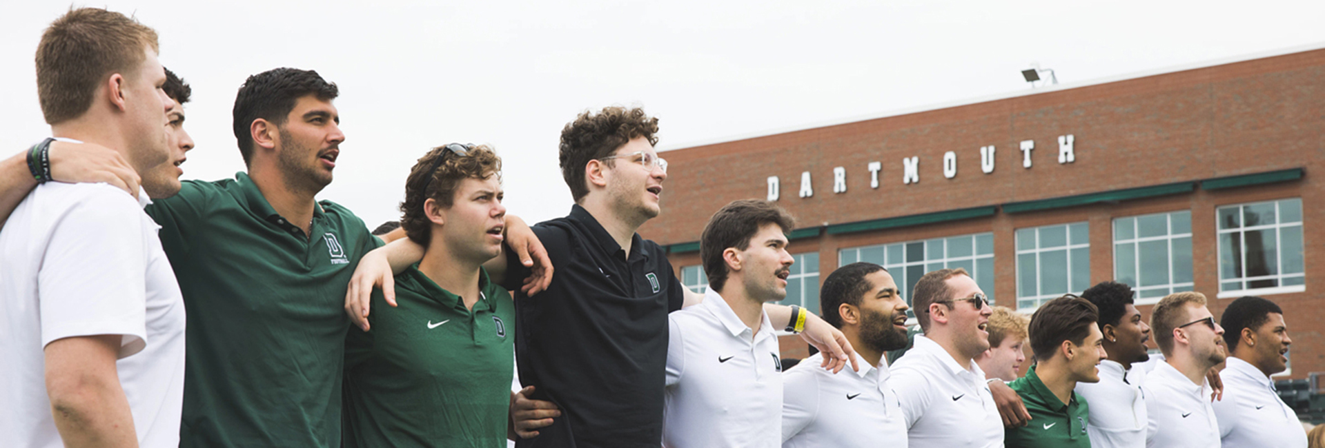 A group of Dartmouth football student athletes standing with their arms on each other's shoulders in song. 