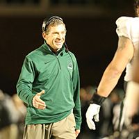 A photo of the late Buddy Teevans on the football field walking toward a student-athlete, smiling, with his hand extended for a hand shake.