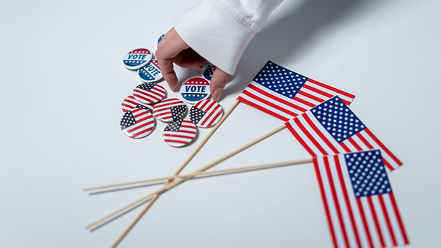 A human hand selecting a button with the word "vote" on it from a pile of US flag buttons next to three small US flags.